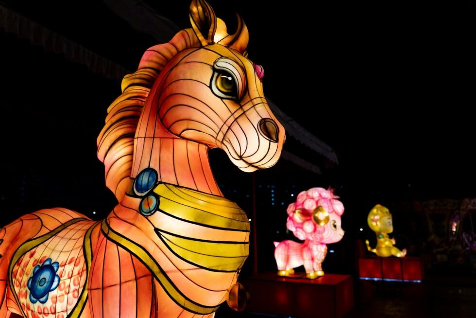 Illuminated lanterns of zodiac animals, featuring a large, detailed horse (2026 zodiac year) in the foreground and vibrant pink and yellow figures in the background, set against a dark night sky.